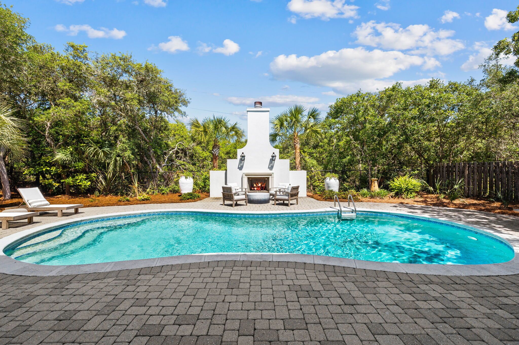 A swimming pool with a white outdoor fireplace, surrounded by lush greenery and a wooden fence.
