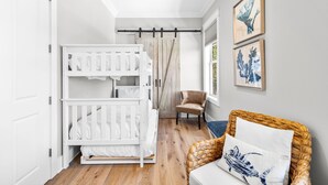 A baby room with a white crib, a wicker chair, and a wall-mounted shelf. - Professionally Decorated with LSV and Pool (Seacrest Beach)