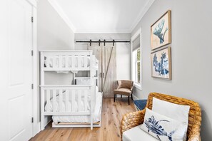 A baby room with a white crib, a wicker chair, and a wall-mounted shelf. - Professionally Decorated with LSV and Pool (Seacrest Beach)