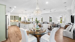 A spacious living area with a glass-top coffee table surrounded by white chairs, a chandelier, and a view of the kitchen in the background. - Professionally Decorated with LSV and Pool (Seacrest Beach)