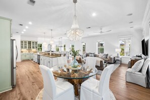 A spacious living area with a glass-top coffee table surrounded by white chairs, a chandelier, and a view of the kitchen in the background. - Professionally Decorated with LSV and Pool (Seacrest Beach)