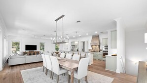 A spacious living room with a large white sofa, a dining area with a wooden table and white chairs, and a kitchen with white cabinets and a stone fireplace. - Professionally Decorated with LSV and Pool (Seacrest Beach)