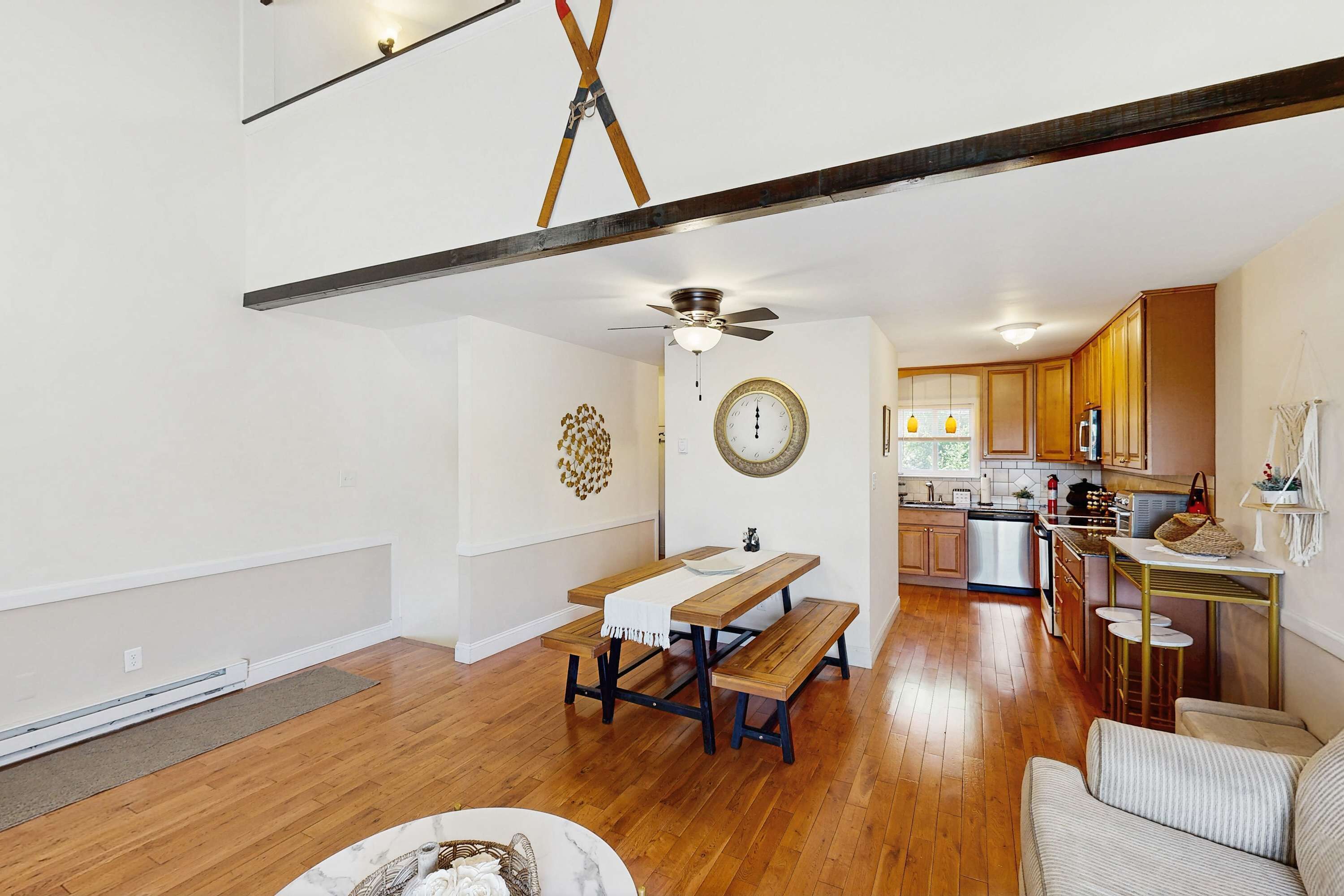 A spacious living area with a wooden floor, a dining table with benches, a ceiling fan, and a kitchen in the background.