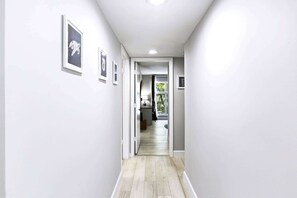 A hallway with light-colored walls and a wooden floor, leading to a room with a window and a door. - Lagoon-View Villa - Walk to Shelter Cove (Hilton Head Island)