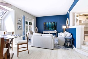A modern living room with a blue accent wall, a large painting, and a dining area with a glass table and chairs. - Lagoon-View Villa - Walk to Shelter Cove (Hilton Head Island)