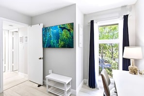 Ett modernt rum med en stor TV, ett fönster med gardiner, ett skrivbord med en lampa och en vit bänk.