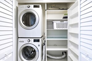 Ett tvättrum med två tvättmaskiner placerade ovanpå varandra, hyllor med tvättkorgar och en ventilationskanal.