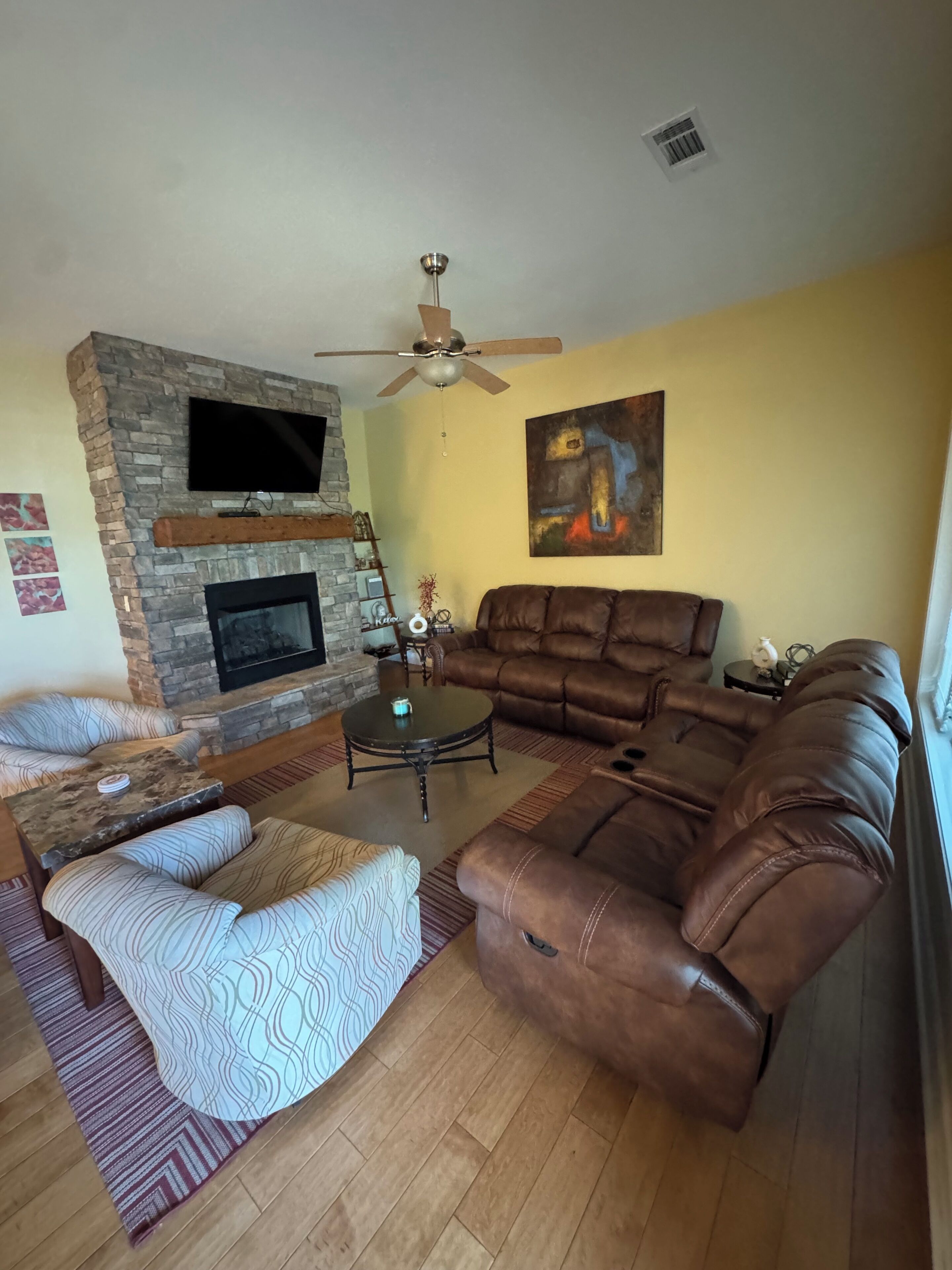 A living room with a stone fireplace, leather sofa, patterned armchair, and a flat-screen TV mounted on the wall.
