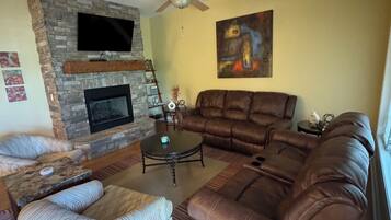 A living room with a stone fireplace, leather sofa, patterned armchair, and a flat-screen TV mounted on the wall.