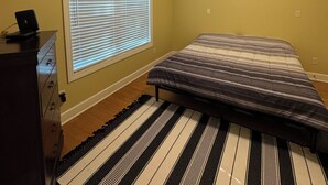 Ein Schlafzimmer mit einem Bett, einem Deckenventilator, einer Kommode, einem Fenster mit Jalousien und einem Streifen Teppich.