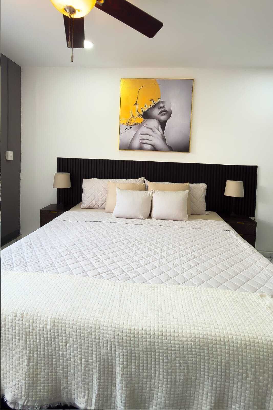 A bedroom with a large bed, two bedside tables, a ceiling fan, and a wall-mounted artwork.