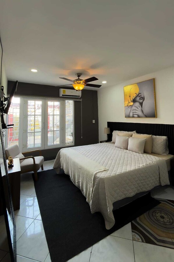 A hotel room with a bed, a desk, a chair, a ceiling fan, and a large window.