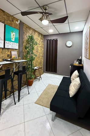 A compact living space with a black sofa, a dining area with chairs, a potted plant, a clock, and a ceiling fan.