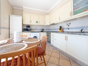A kitchen with a dining area, including a table with placemats, chairs, a microwave, a coffee maker, and a washing machine. - Quinta Gomeira - Top Floor 2 Bedroom with pool (Cabanas de Tavira)