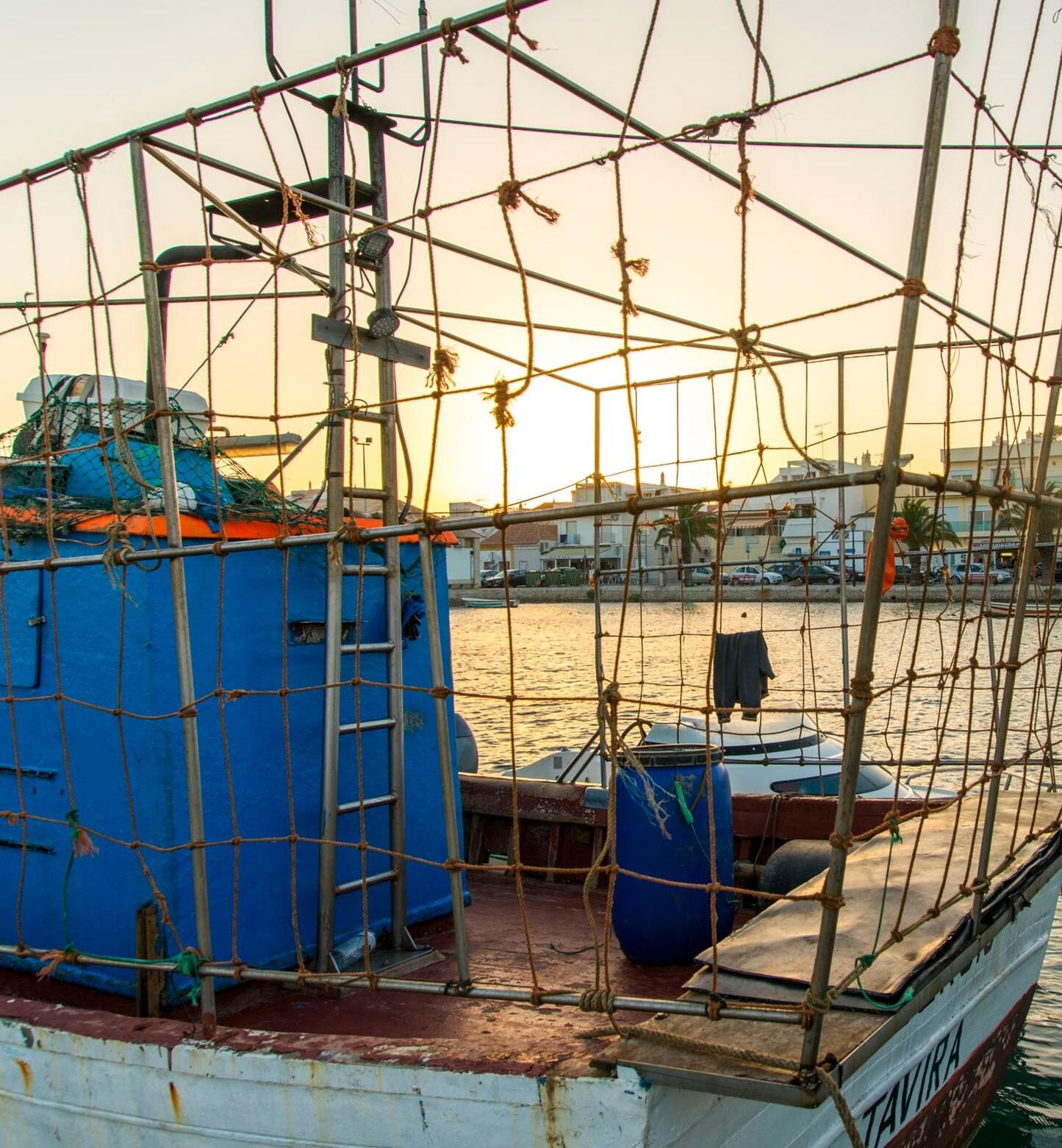 Ein Boot mit einer blauen Kabine und einem Netzabdecknetz, angedockt in einem Hafen, mit Gebäuden im Hintergrund.
