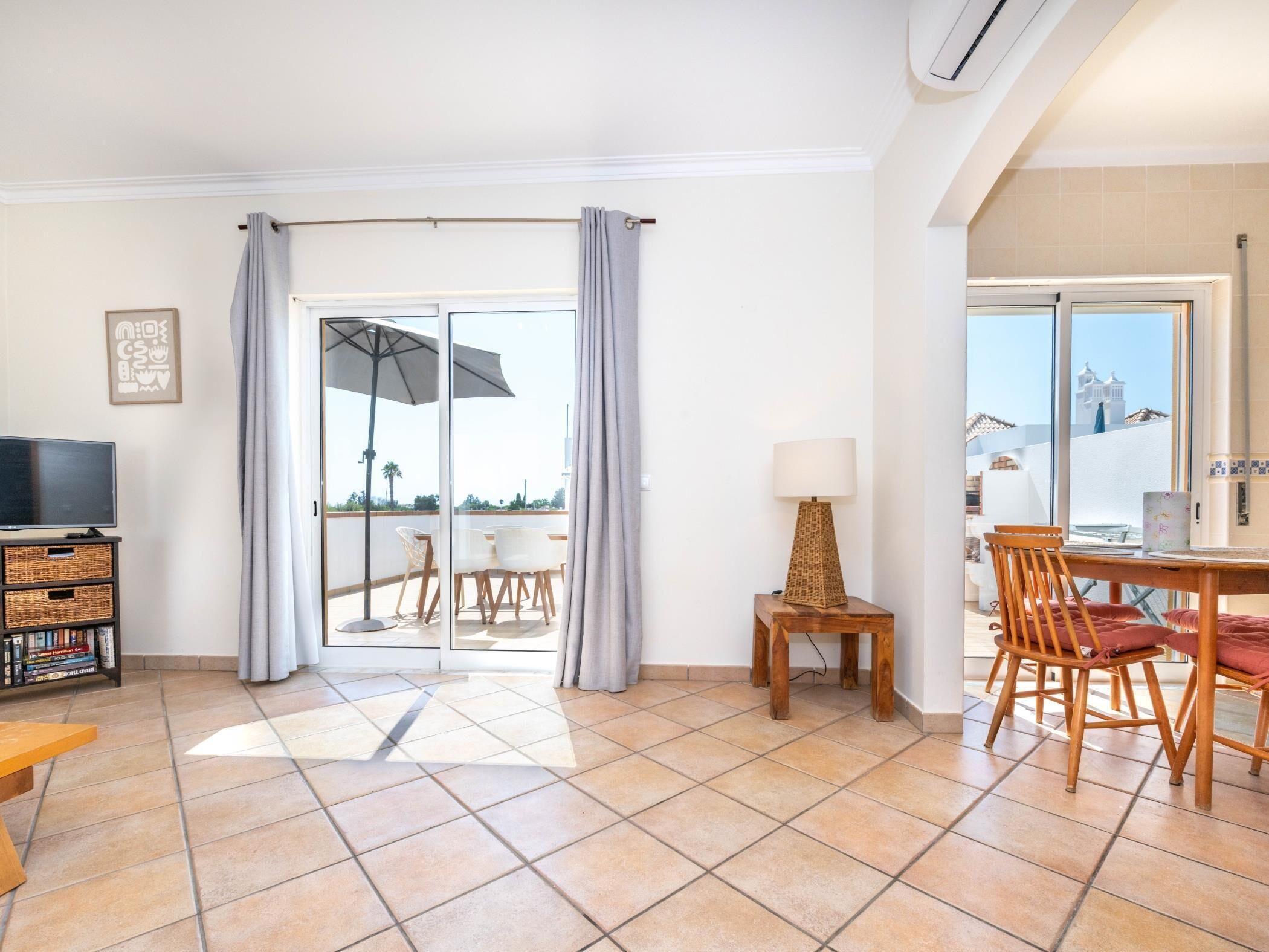 Une pièce spacieuse avec un balcon, une télévision, une table à manger avec des chaises et une lampe.
