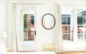 A room with a bed, a round mirror, a bench, and a view of a brick building through a glass door. - Park House Parlor Level - 2 Bedroom/2 Bath Overlooking Monterey Square (Savannah)