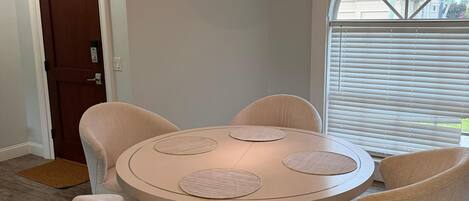Een eetruimte met een ronde tafel, zes stoelen, een hanglamp, een deur en een raam met zonwering.