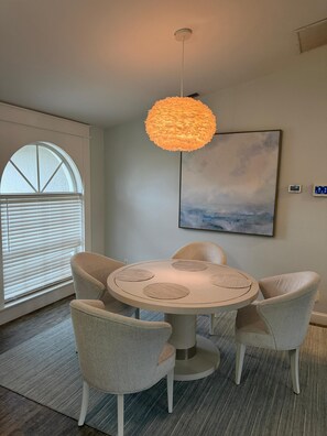 Uma área de refeições com uma mesa redonda, quatro cadeiras, um lustre, uma janela com persianas e um quadro na parede.