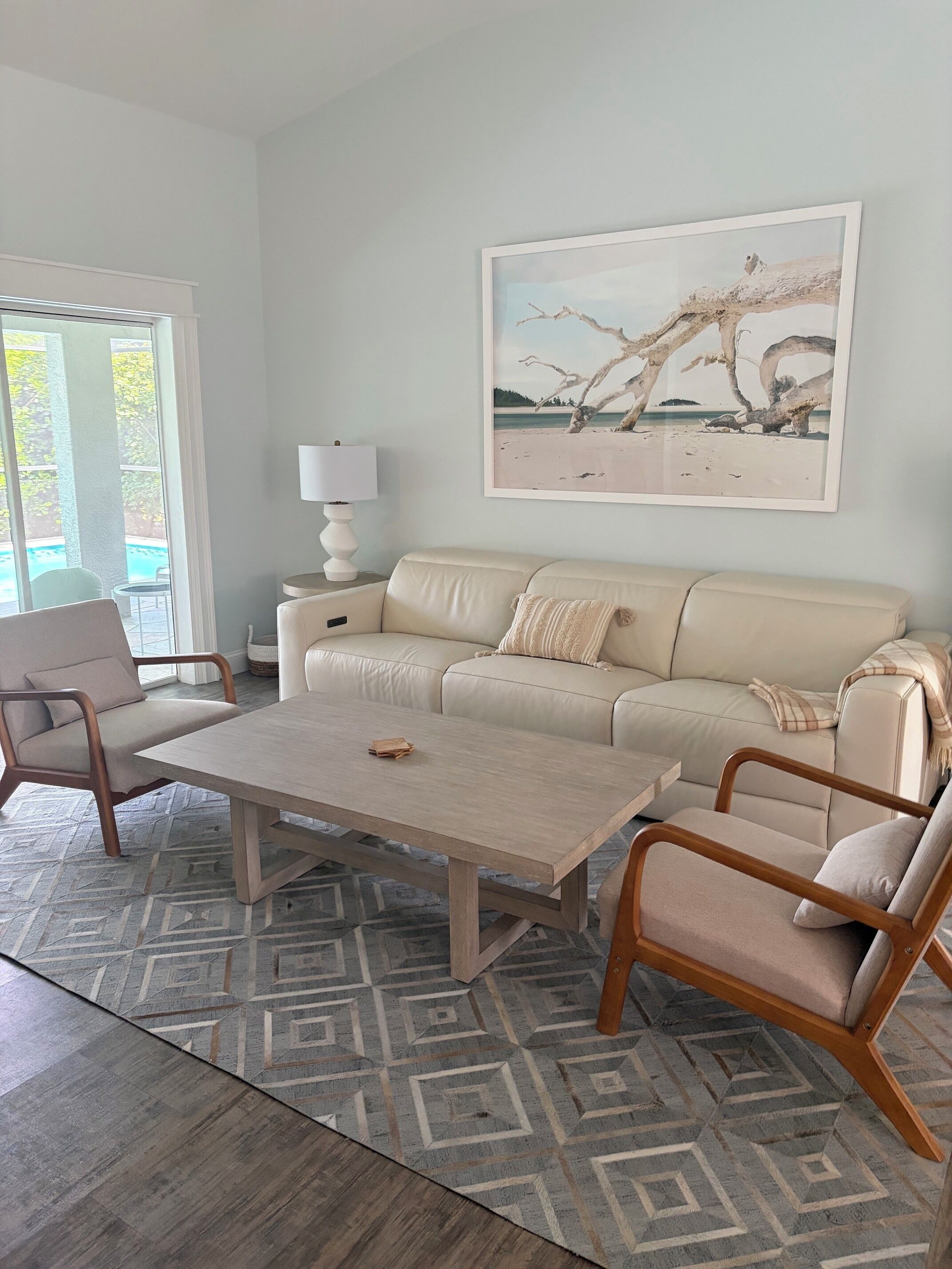 A modern living room with a beige sofa, two armchairs, a coffee table, and a large painting of a beach scene.