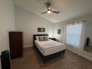 Une chambre à coucher comprenant un lit, un ventilateur de plafond, une table de chevet, une commode et une fenêtre avec des stores.