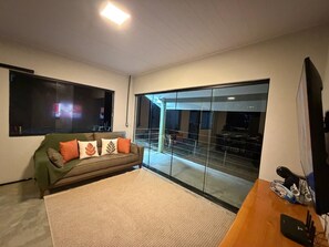 A living room with a sofa, a flat-screen TV, a wooden coffee table, and a large glass door leading to an outdoor area. - Deusmideu Refuge (São Bento do Sapucaí)