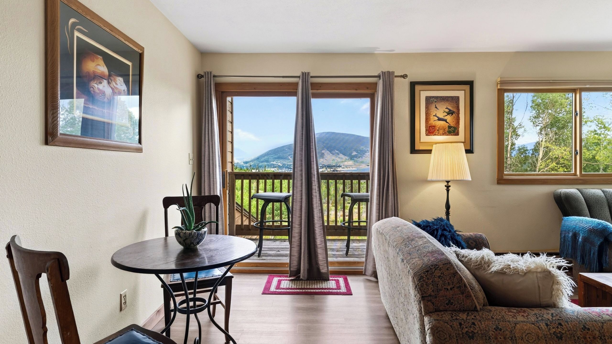 A room with a dining table, chairs, a sofa, a lamp, and a view of a mountain and lake through a sliding door.