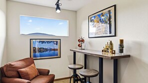 Ein Zimmer mit einer braunen Ledersofa, einem Couchtisch mit Metallgestell und zwei Barhocker. An der Wand hängen drei gerahmte Bilder, eine Deckenleuchte und auf dem Tisch steht eine Flasche.