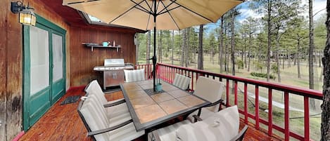 A wooden deck with a table and chairs, a grill, and a view of the surrounding forest.
