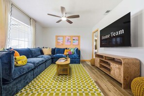 A living room with a blue sofa, a wooden coffee table, a checkered floor, a ceiling fan, and a TV on the wall. - Pool •  Hot Tub•  Putt Putt • Game Room •  Karaoke (Largo)