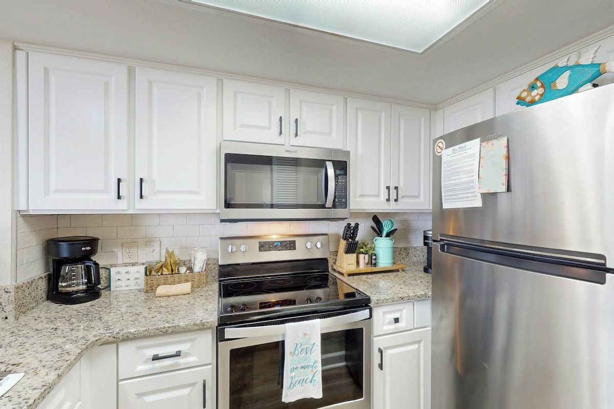 Una cocina con gabinetes blancos, un refrigerador de acero inoxidable, un microondas, un horno y una cafetera.