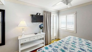 Una camera da letto con un letto, un comodino bianco, una lampada, una televisione a parete, una ventola a soffitto e una finestra con persiane.