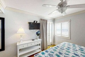 Ein Schlafzimmer mit einem Bett, einem weißen Nachttisch, einer Lampe, einem an der Wand befestigten Fernseher, einem Deckenventilator und einem Fenster mit Jalousien.