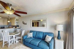 A living room with a blue sofa, a dining table with chairs, a ceiling fan, and a kitchen area in the background. - A Place at the Beach V - A307 (Myrtle Beach)