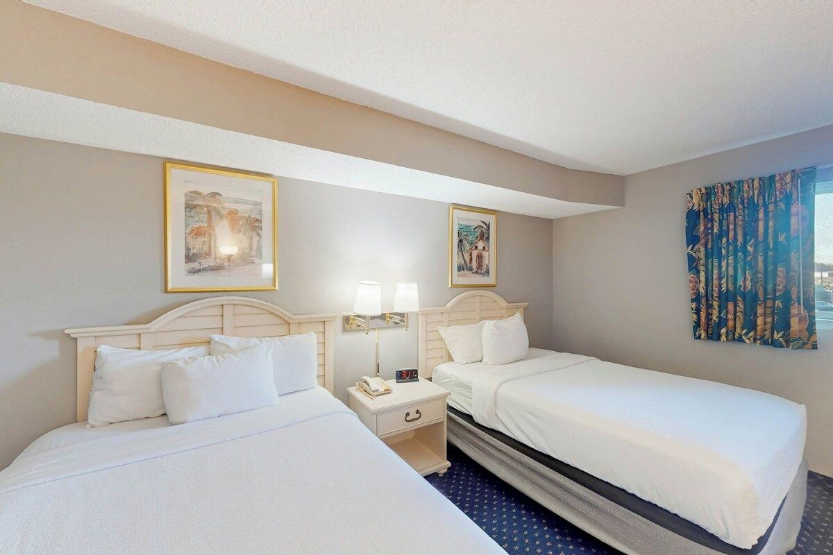 Habitación de hotel con dos camas, una mesita de noche con lámpara y una ventana con cortinas.