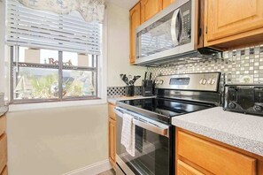 A kitchen with wooden cabinets, a microwave, a stove, and a window with blinds. - Arcadian Dunes 02-207 (Myrtle Beach)