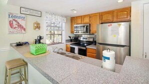 A kitchen with granite countertops, wooden cabinets, stainless steel appliances, and a green stool. - Arcadian Dunes 02-207 (Myrtle Beach)