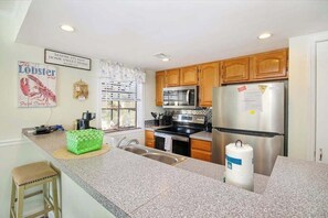 A kitchen with granite countertops, wooden cabinets, stainless steel appliances, and a green stool. - Arcadian Dunes 02-207 (Myrtle Beach)