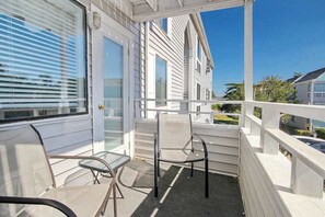 A covered porch with a glass table and two chairs, a white railing, and a view of a residential street. - Arcadian Dunes 02-207 (Myrtle Beach)