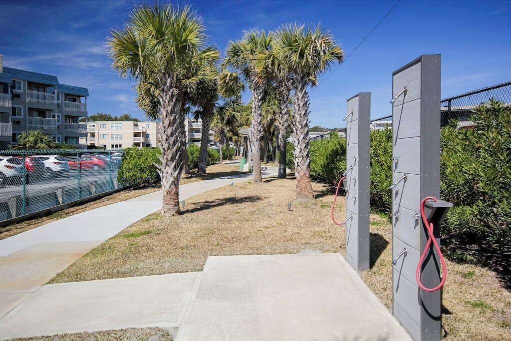 A parking area with a concrete pathway leading to a gated entrance, palm trees, and a fence surrounding the area.
