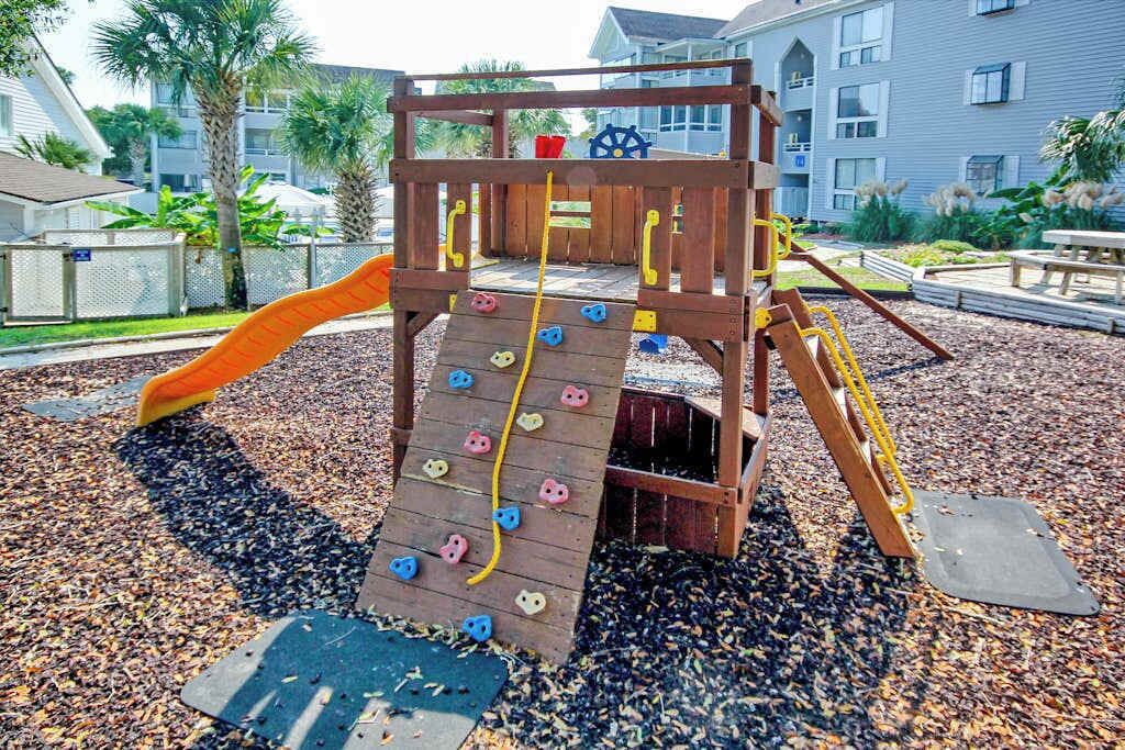 A wooden play structure with a slide and climbing wall, surrounded by a playground area with mulch ground cover.