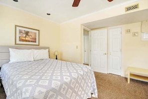 A bedroom with a bed, a bench, a door, and a wall-mounted picture. - Arcadian Dunes 02-207 (Myrtle Beach)