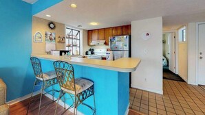 A kitchen with a blue accent wall, wooden cabinets, a refrigerator, and two bar stools. - Arcadian Dunes 02-208 (Myrtle Beach)