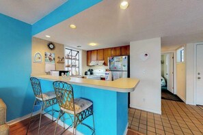 A kitchen with a blue accent wall, wooden cabinets, a refrigerator, and two bar stools. - Arcadian Dunes 02-208 (Myrtle Beach)