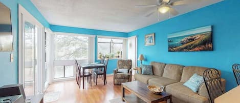 Ein Wohnzimmer mit blauer Wand, Deckenventilator, einer Couch, einem Couchtisch und einem Essbereich mit Stühlen und einem Tisch.