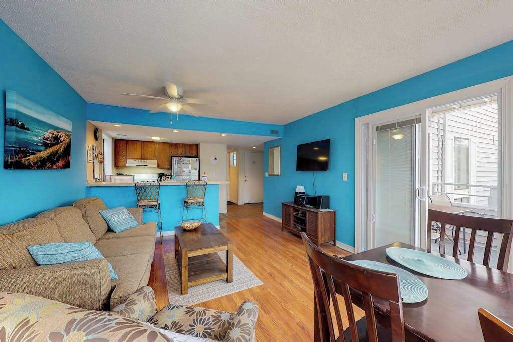 A living room with a sofa, coffee table, and TV. There's a kitchen area with cabinets and a refrigerator.