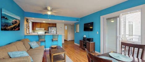 A living room with a sofa, coffee table, and TV. There's a kitchen area with cabinets and a refrigerator.