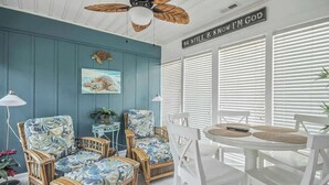 A room with a ceiling fan, a wall with a sign, a table with chairs, and a window with blinds. - Cozy Beach House 314 (North Myrtle Beach)