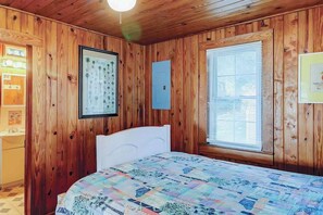 A wooden-paneled room with a bed, a framed picture of palm trees, a window with blinds, and a small kitchenette. - Ocean Gem (North Myrtle Beach)