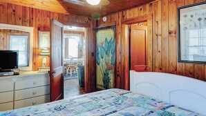 Ein Zimmer mit Holzwandverkleidung, einem Bett, einem Fernseher, einem Schreibtisch mit Lampe und einer Tür zu einem anderen Raum.